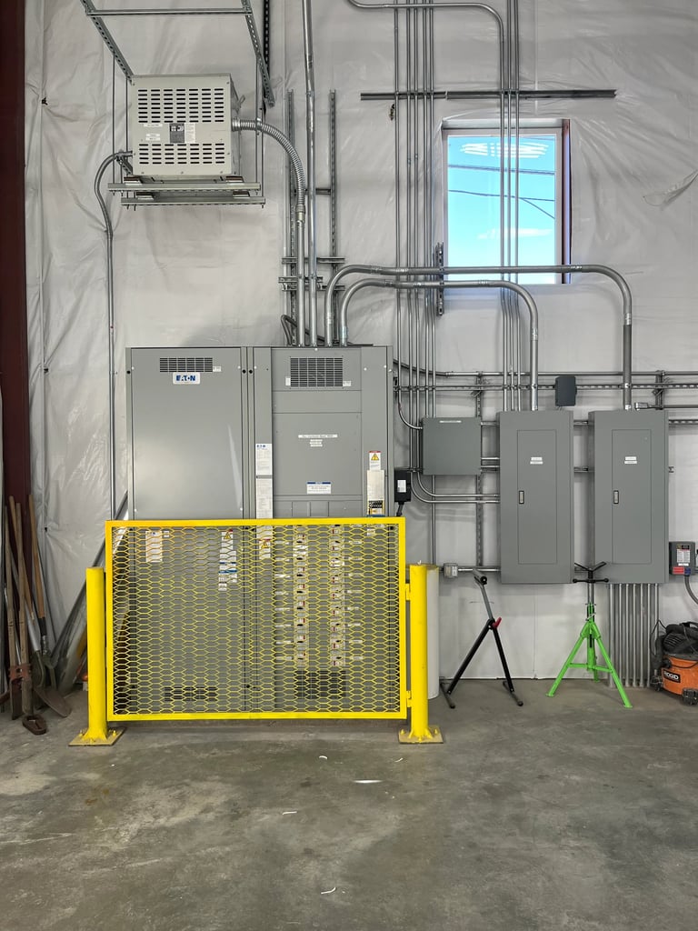 Industrial electrical room with yellow mesh safety gate, gray electrical panels, conduit piping, and a blue window in the background